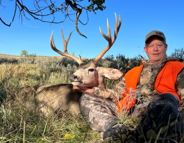 A nonresident Wyoming hunter poses with his mule deer amongst the sagebrush in his camo clothing and orange vest